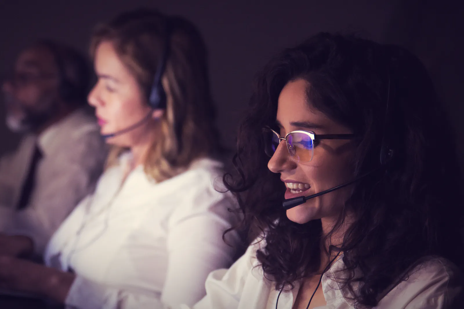 Vendedora sorrindo com fone de ouvido durante uma ligação, com colegas ao fundo.